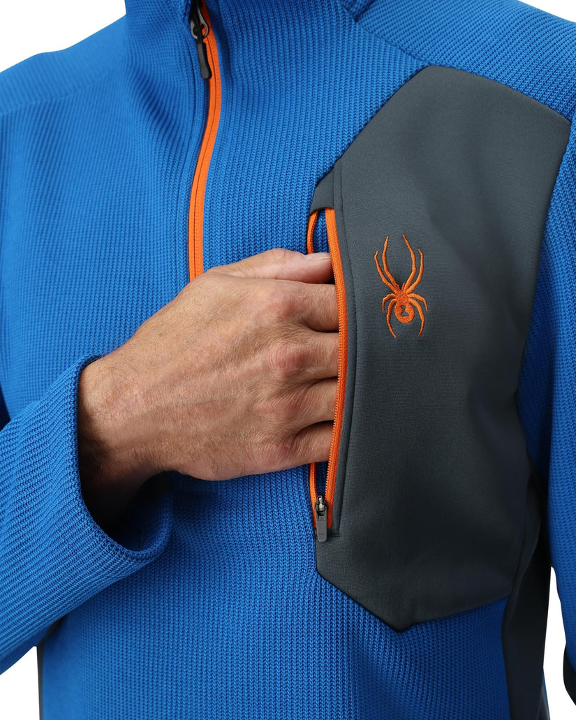 Blue jacket with orange zipper and black pouch featuring a logo, held by a hand.