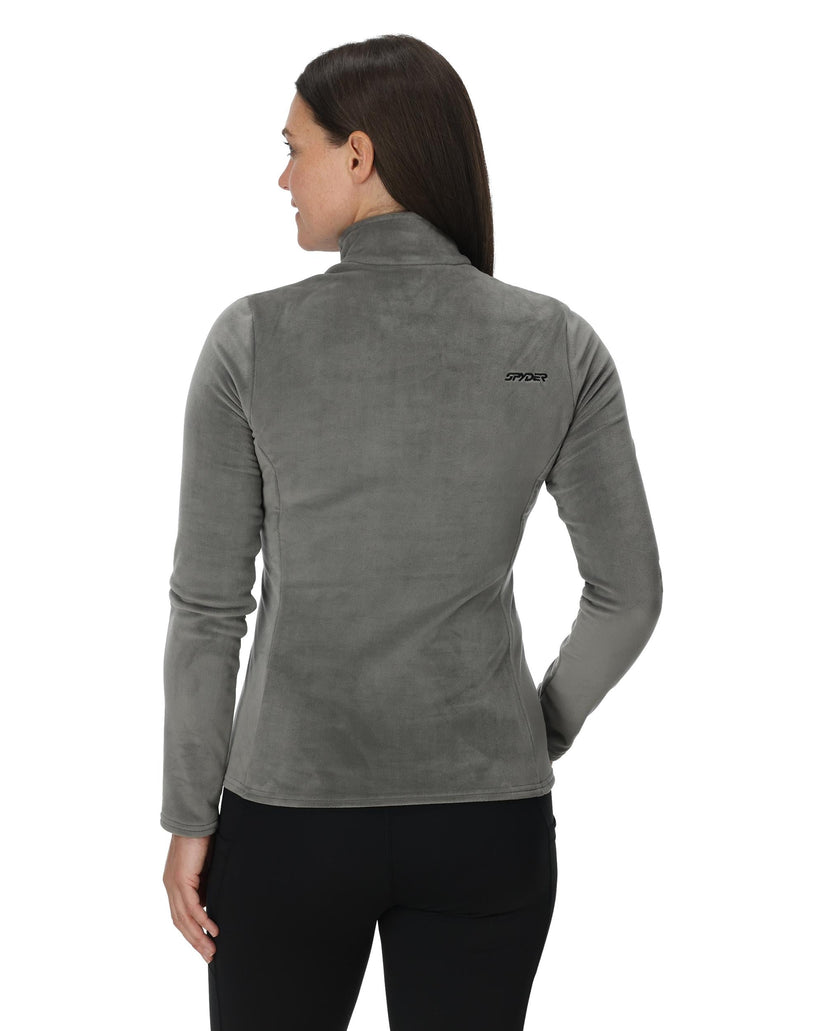 Woman wearing a gray fleece jacket with a brand logo on the back, standing against a white background.