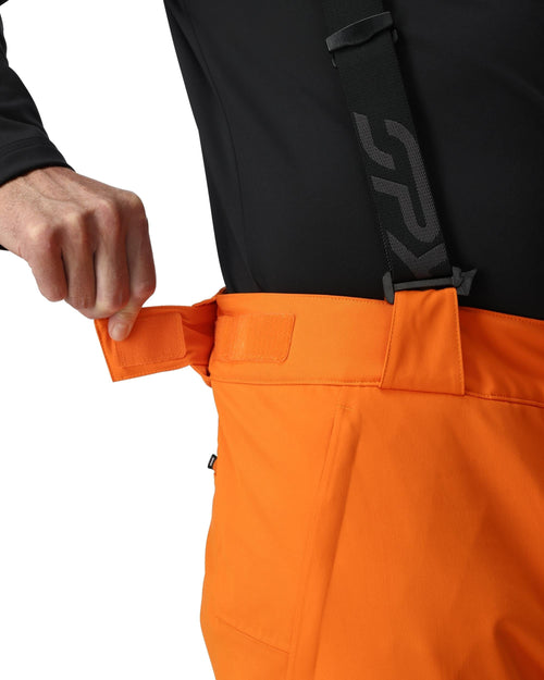 Man wearing orange snow pants and black top with a brand logo on a white background